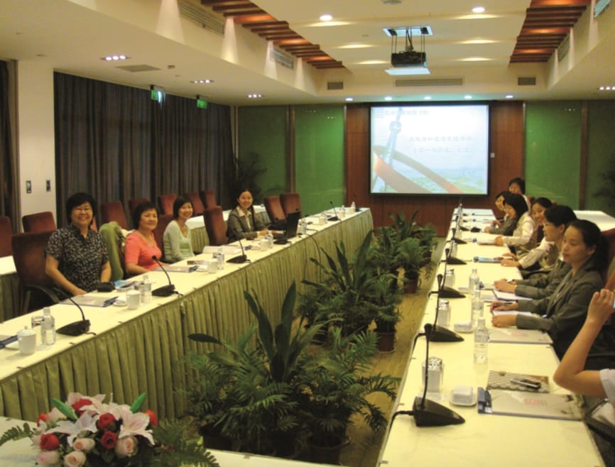 A conference room with people seated around a U-shaped table, microphones, documents, and a projection screen displaying a presentation.